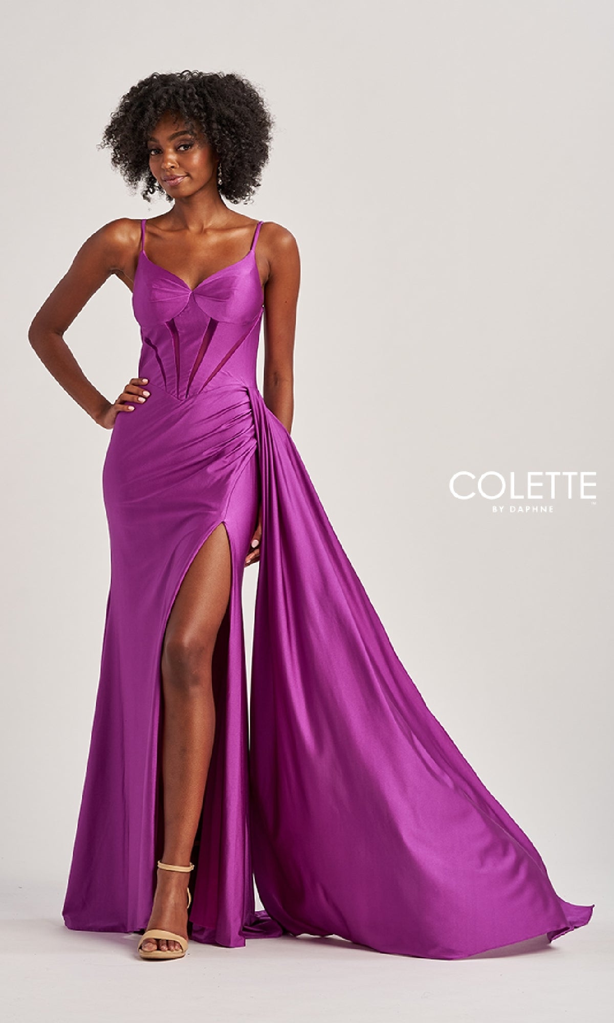 A woman wears the Sheer-Cut-Out Long Prom Dress: Colette CL8480 in bright purple, featuring a lace-up bodice, thigh-high slit, and draped fabric as she poses against a neutral background.